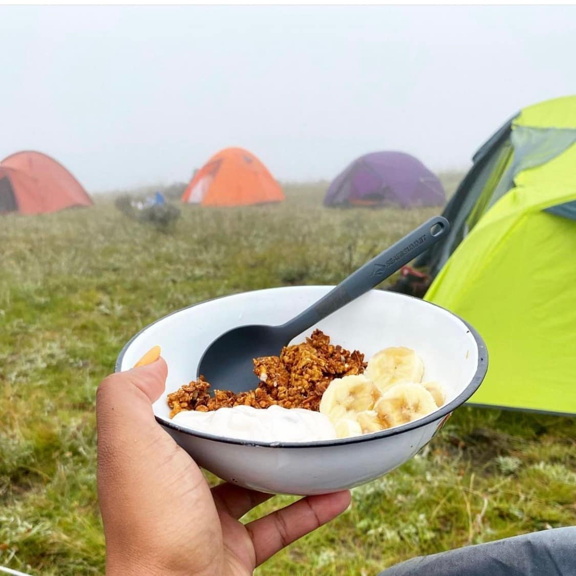 Bowl of Hungry Hiker gluten-free granola with yogurt and banana at a campsite with tents in the background
