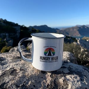 Hungry Hiker white enamel camping mug with black rim displayed on a rock overlooking a mountain landscape.
